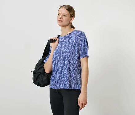 Frau mit schwarzem Rucksack über der Schulter trägt ein blau gemustertes T-Shirt und schwarze Leggings.
