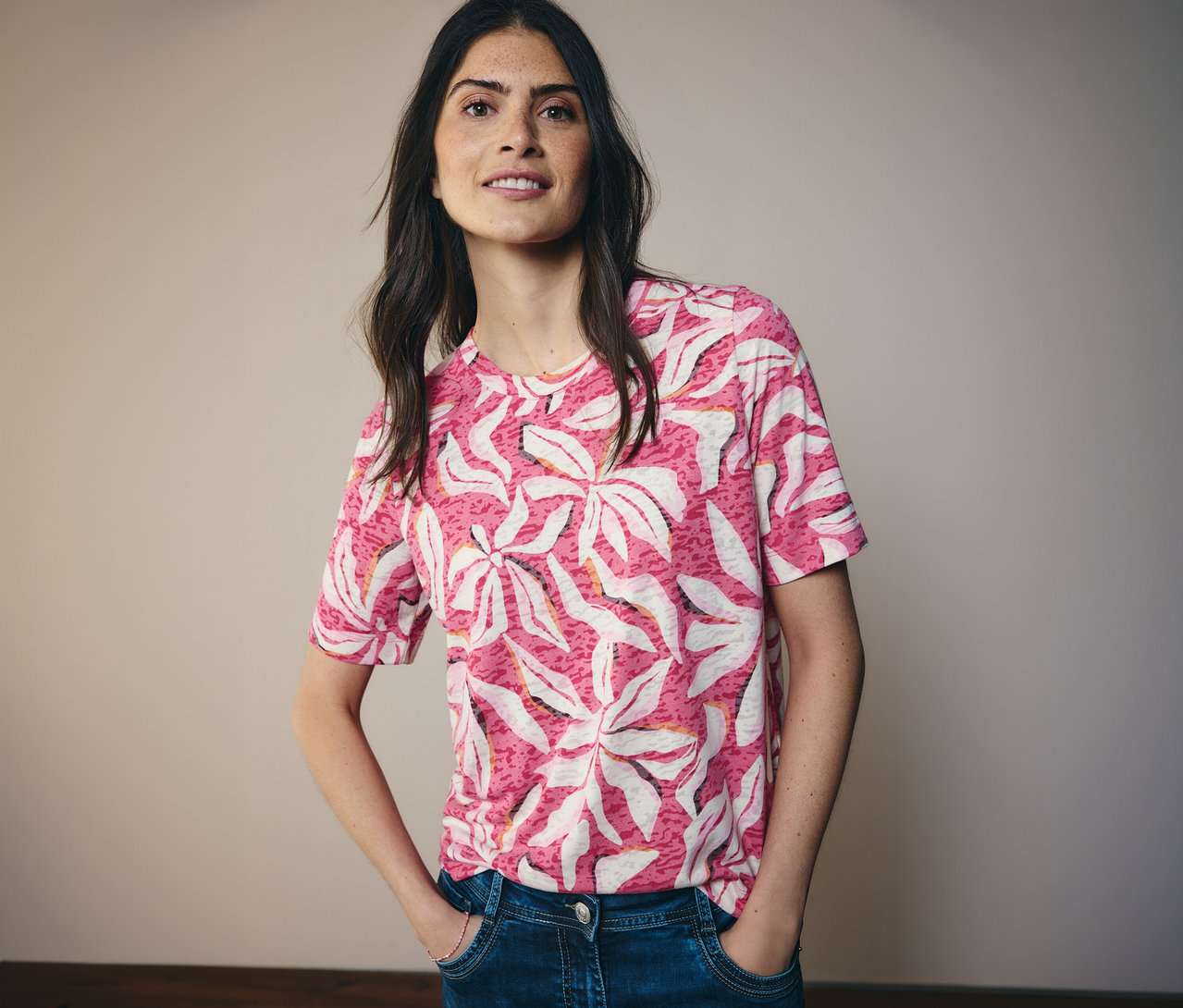 Frau mit Jeans und rosa CECIL Kurzarmshirt mit Blätterprint. Sie steht vor einer hellen Wand.