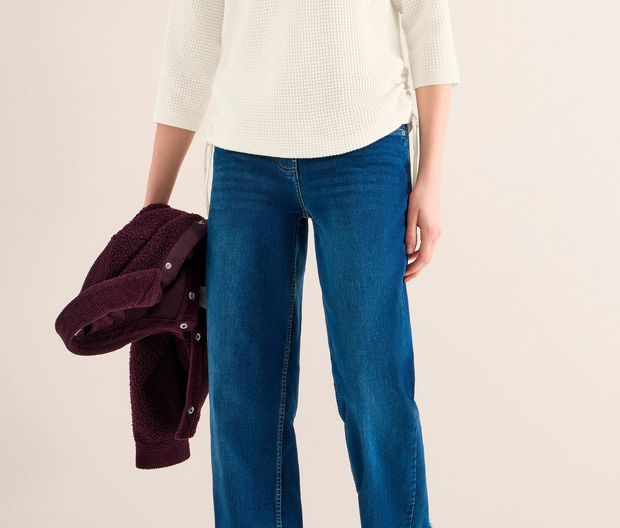 Frau trägt einen weißen CECIL Feinstrickpullover, blaue Jeans mit weitem Bein und Krempelbund sowie Sneakers und hält eine bordeauxfarbene Jacke.