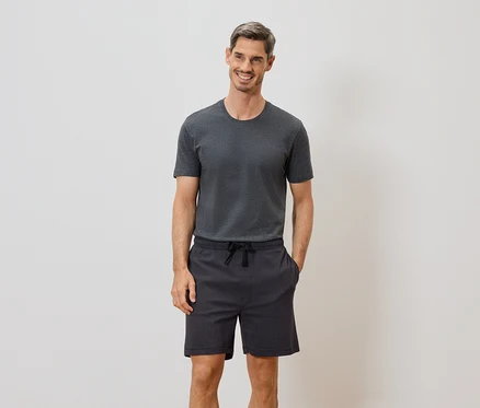 Mann mit grauem T-Shirt und Shorts mit Kordelzug steht vor einer weißen Wand und lächelt.