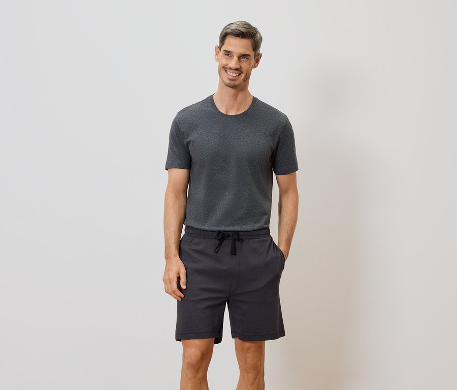 Mann mit grauem T-Shirt und Shorts mit Kordelzug steht vor einer weißen Wand und lächelt.