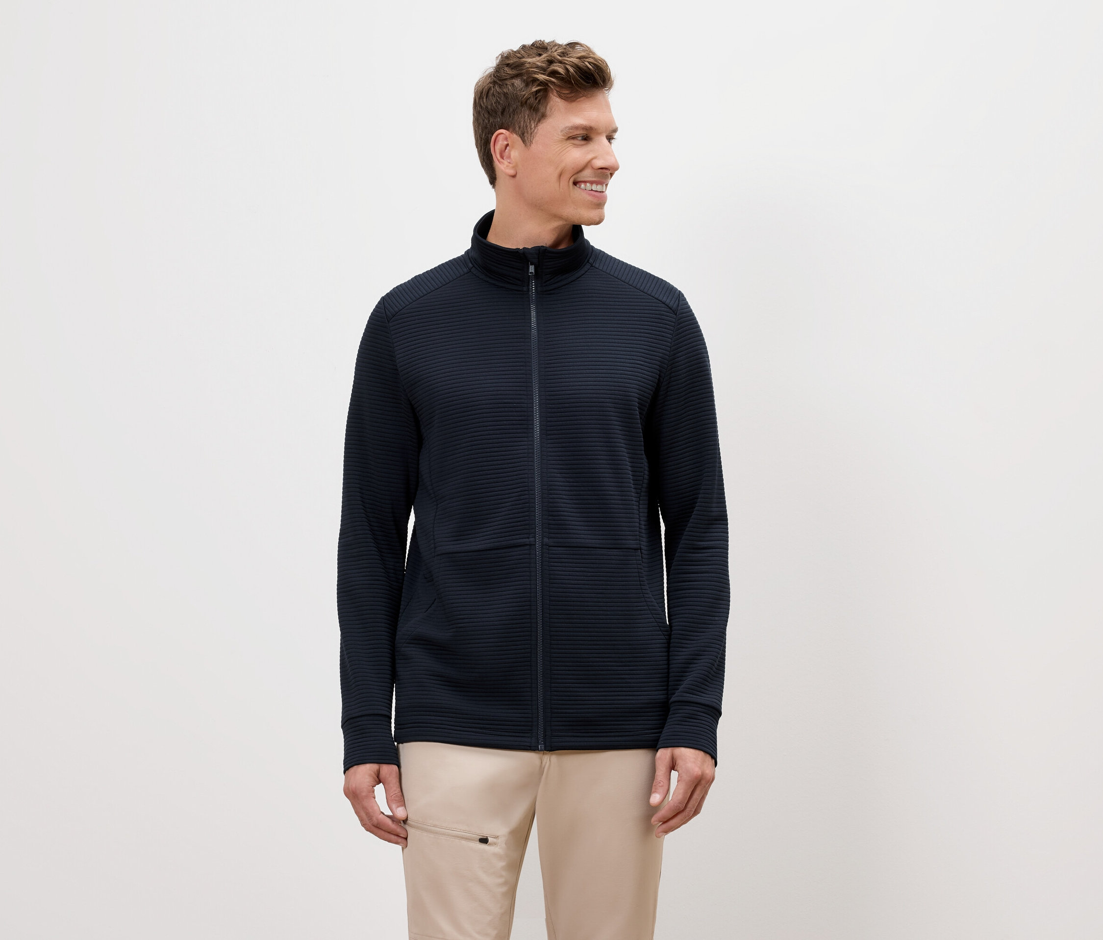 Ein Mann mit blonden Haaren trägt eine marineblaue Reißverschlussjacke und beigefarbene Hosen.