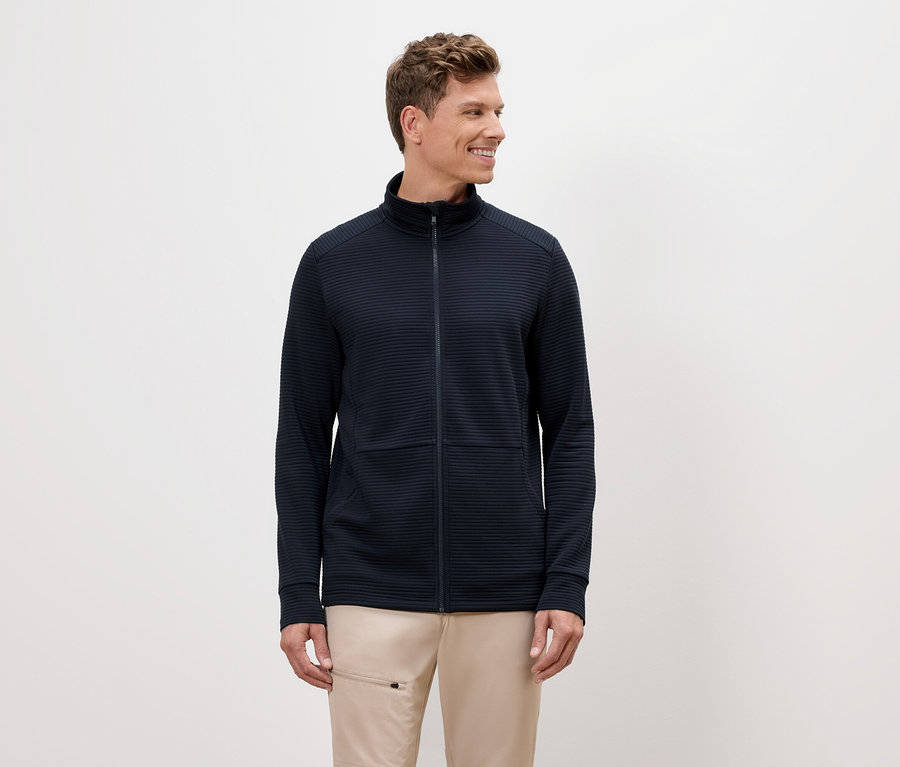 Ein Mann mit blonden Haaren trägt eine marineblaue Reißverschlussjacke und beigefarbene Hosen.