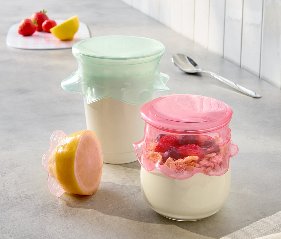 Auf einer Küchentheke stehen zwei Joghurtgläser mit rosa und mintfarbenen Deckeln und eine halbe Zitrone mit einem rosa Deckel.