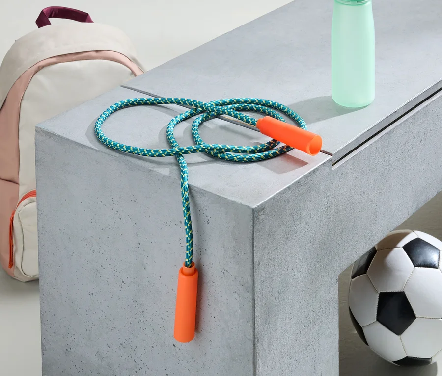 Ein rosa-weißer Rucksack, ein Springseil, eine Trinkflasche und ein Fußball liegen auf einer grauen Bank.