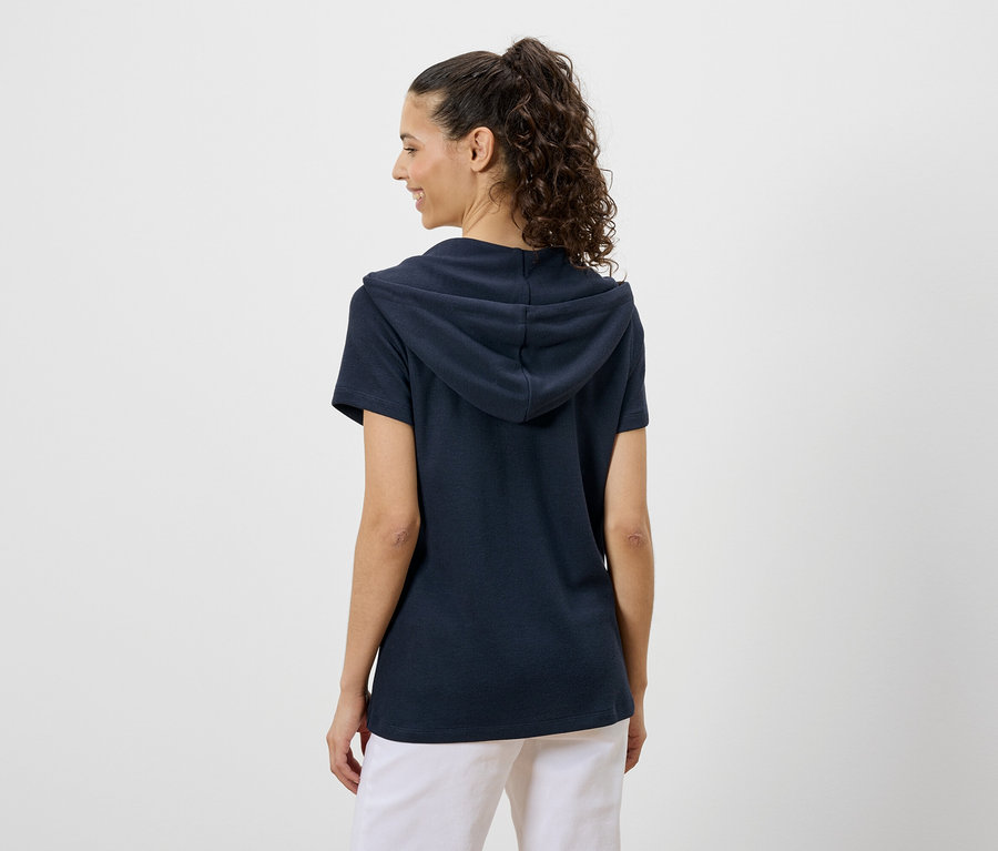 Eine Frau mit lockigem Haar trägt ein marineblaues Kapuzen-T-Shirt und weiße Hosen, von hinten gesehen.