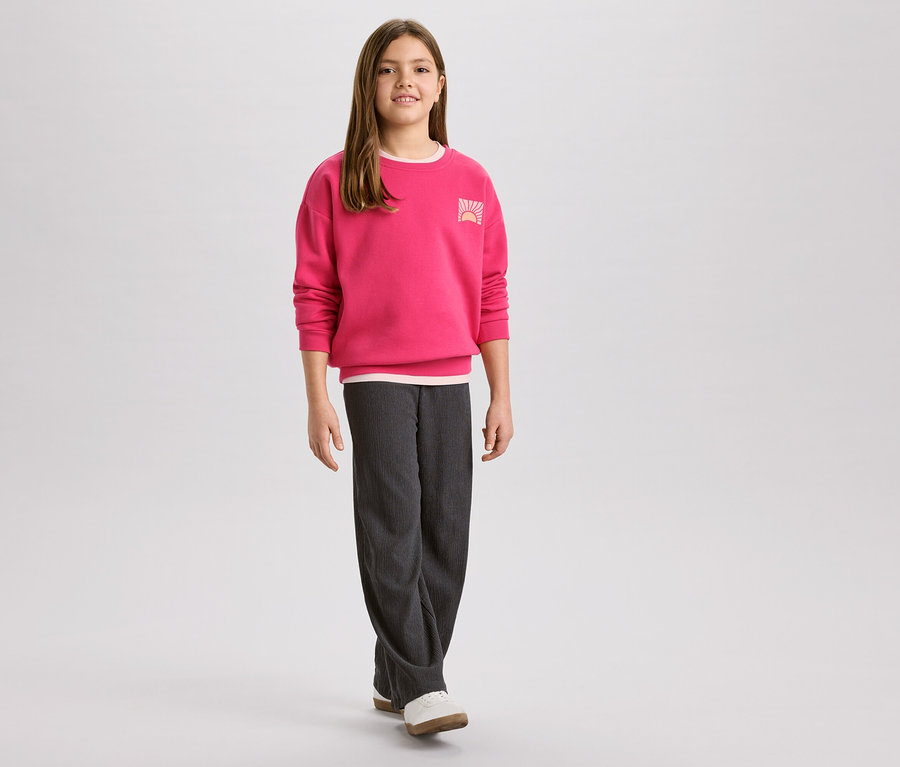 Mädchen mit pinkem Pullover mit Sonnenaufgangslogo und grauer Cordhose, dazu weiße Schuhe.