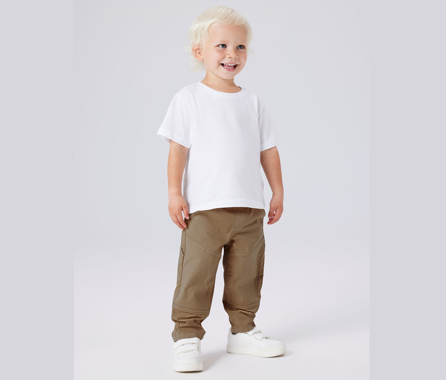 Ein lächelnder, blonder, kleiner Junge steht in einem weißen T-Shirt, Khaki-Hosen und weißen Turnschuhen vor einem grauen Hintergrund.