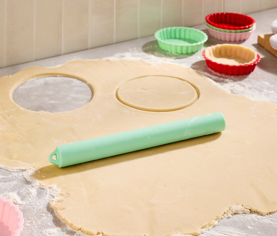 Auf einer bemehlten Arbeitsfläche liegt ausgerollter Teig mit zwei ausgestochenen Kreisen, ein grüner Teigroller und bunte Tarteförmchen.