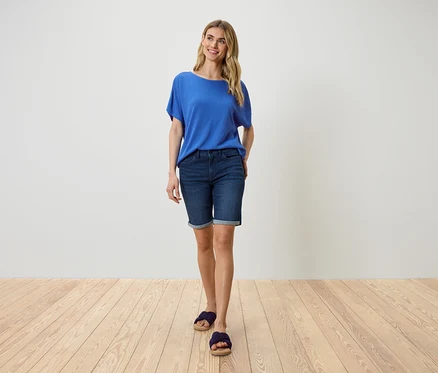 Eine Frau steht vor einer weißen Wand und trägt ein blaues T-Shirt, Jeansshorts und Sandalen.
