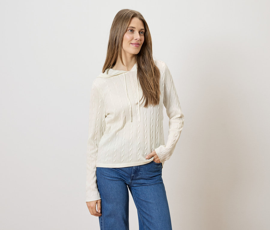 Frau mit langem, braunem Haar trägt einen cremefarbenen Zopfmuster-Hoodie und blaue Jeans.