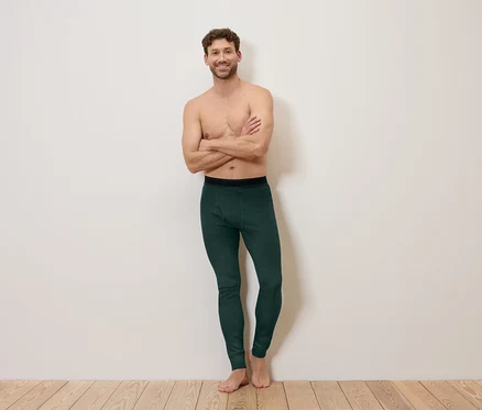 Mann mit verschränkten Armen, barfuß, trägt dunkelgrüne Leggings vor einer weißen Wand.