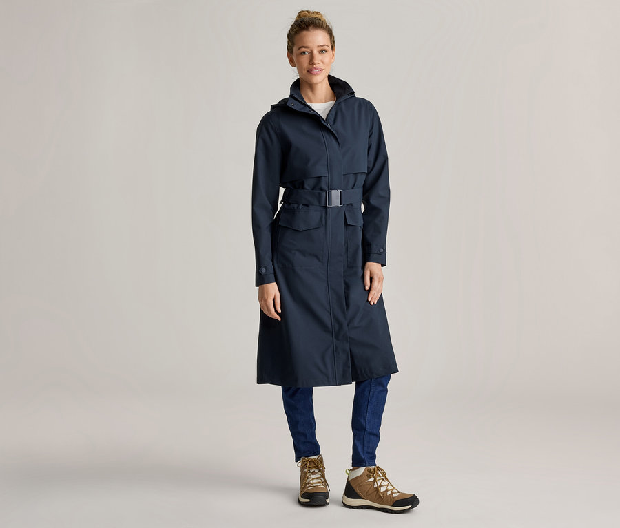 Frau mit Dutt trägt einen langen, marineblauen Regenmantel mit Gürtel, blaue Jeans und braune Wanderschuhe vor einem hellgrauen Hintergrund.