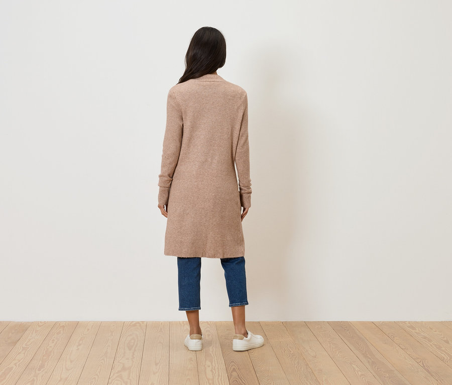Rückansicht einer Frau mit langen, dunklen Haaren, die einen langen, beigen Cardigan, blaue Jeans und weiße Schuhe trägt.