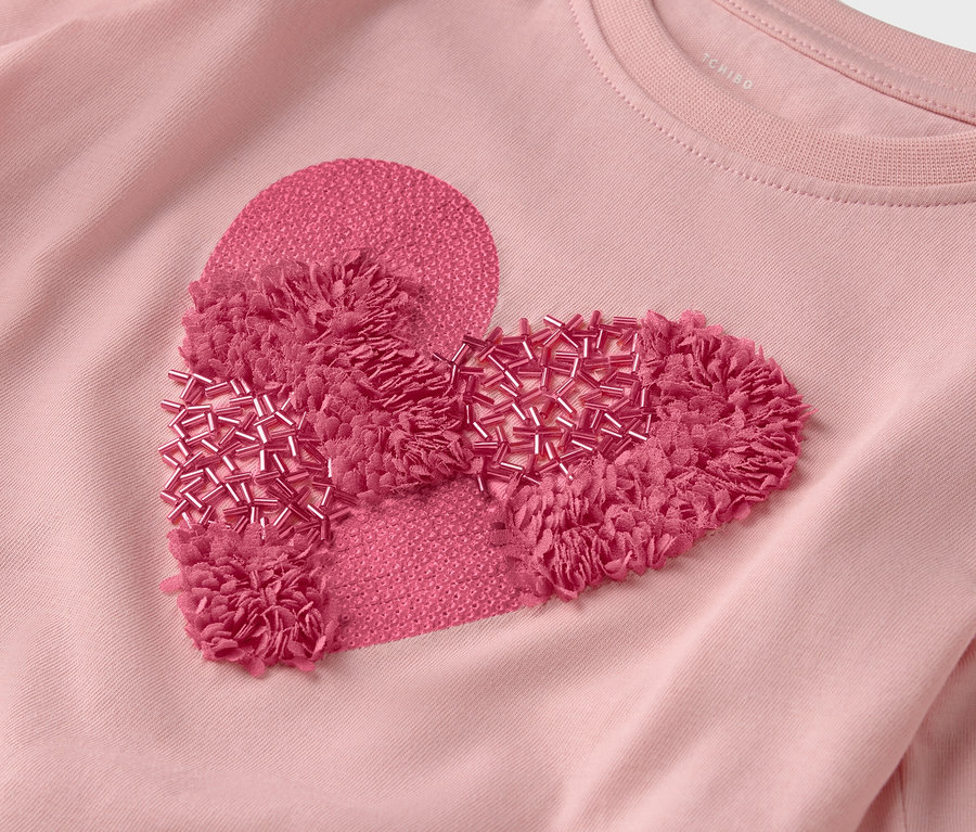 Detailansicht eines rosafarbenen T-Shirts mit einem herzförmigen Aufnäher, der mit Fransen und Perlen verziert ist.