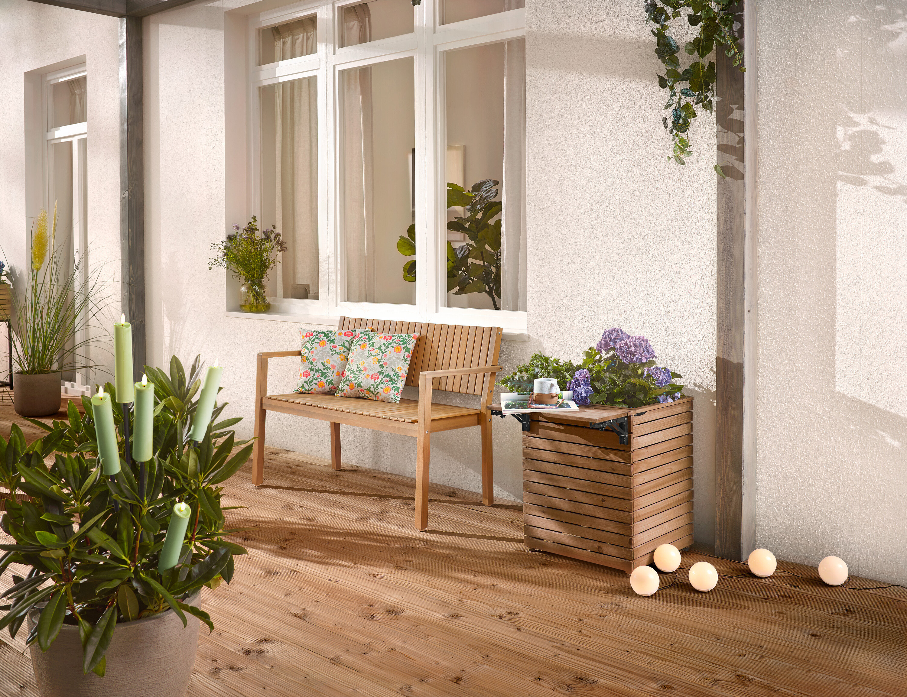 Auf einer Holzterrasse stehen eine Holzbank mit Kissen, ein Pflanzkasten mit Blumen und eine Topfpflanze mit Kerzen.