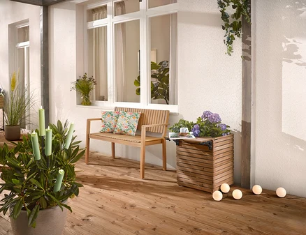 Auf einer Holzterrasse stehen eine Holzbank mit Kissen, ein Pflanzkasten mit Blumen und eine Topfpflanze mit Kerzen.