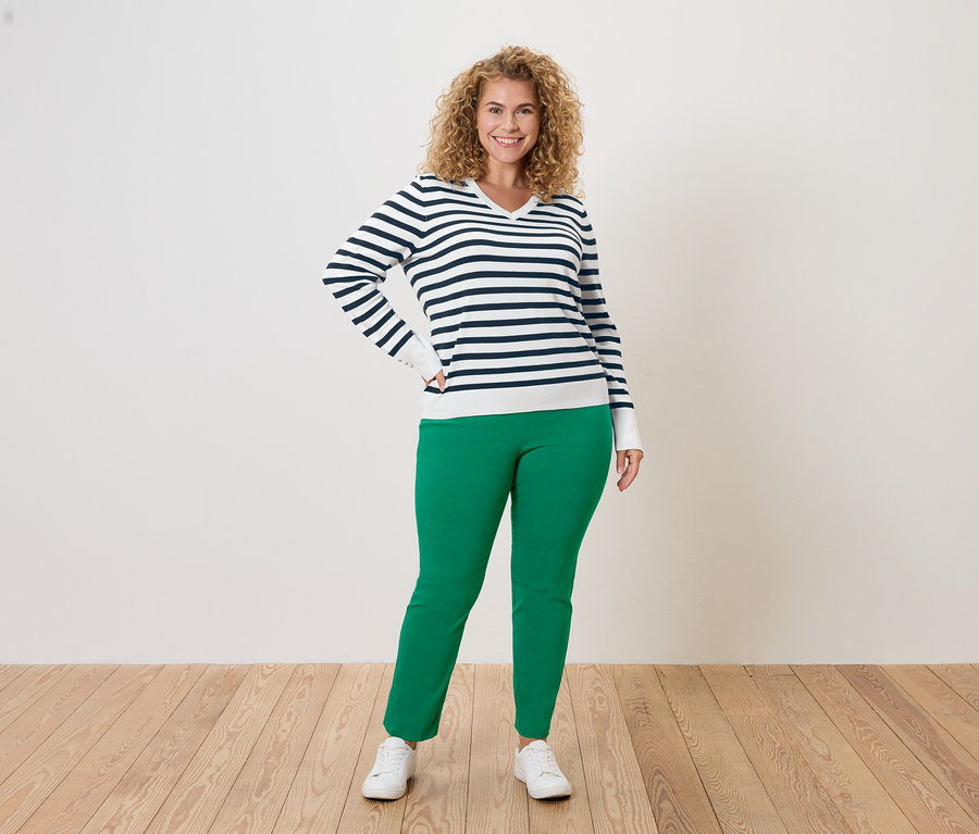 Frau mit lockigem Haar, trägt einen blau-weiß gestreiften Pullover, grüne Hose und weiße Turnschuhe, posiert vor einem weißen Hintergrund.