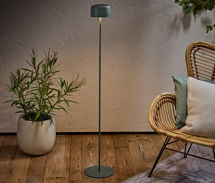 Eine grüne Stehlampe steht neben einem Rattansessel mit Kissen und einer Topfpflanze auf einem Holzboden.