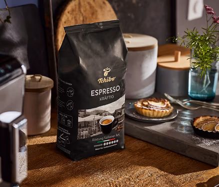 Eine Packung Tchibo Espresso Kräftig Ganze Bohne steht auf einem Holztisch neben einer Kaffeemaschine und Tellern mit Törtchen.