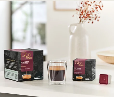 Qbo Espresso APANECA ANA Packungen mit 27 und 8 Kapseln neben einem Glas Espresso und einem einzelnen Kapselwürfel.