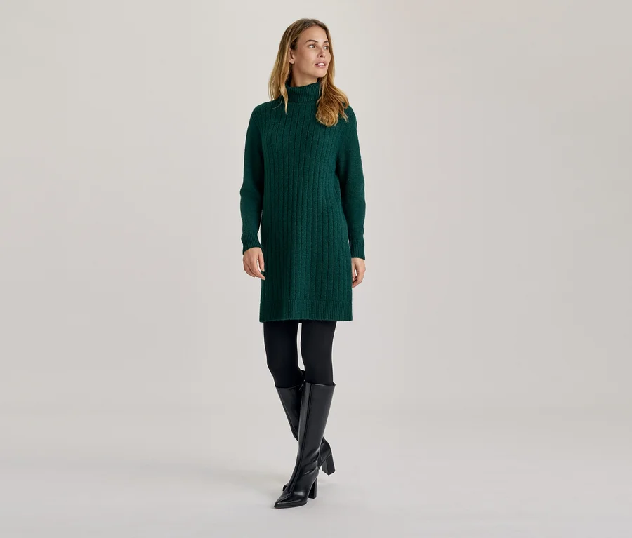 Frau in einem grünen Rollkragen-Strickkleid, schwarzen Leggings und schwarzen kniehohen Stiefeln, die zur Seite blickt.