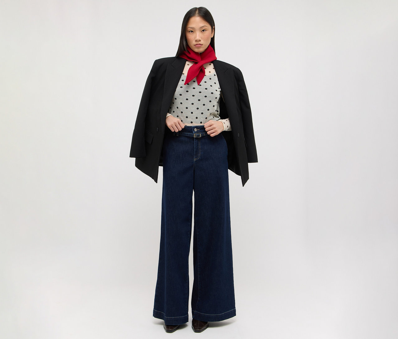 Eine Frau steht da und trägt einen schwarzen Blazer über den Schultern, ein helles gepunktetes Oberteil, dunkelblaue weite Jeans und einen roten Schal um den Hals.