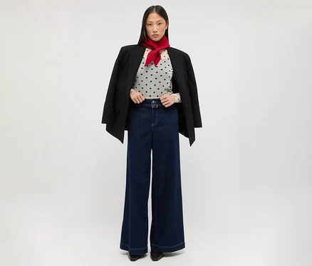 Eine Frau steht da und trägt einen schwarzen Blazer über den Schultern, ein helles gepunktetes Oberteil, dunkelblaue weite Jeans und einen roten Schal um den Hals.