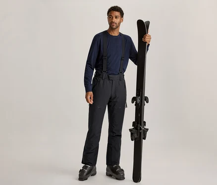 Ein Mann mit dunklem Lockenhaar und hellem Bart steht da, trägt ein dunkelblaues Langarmshirt, schwarze Skihosen mit Hosenträgern und schwarze Skischuhe. Er hält ein Paar schwarze Skier aufrecht in seiner rechten Hand.