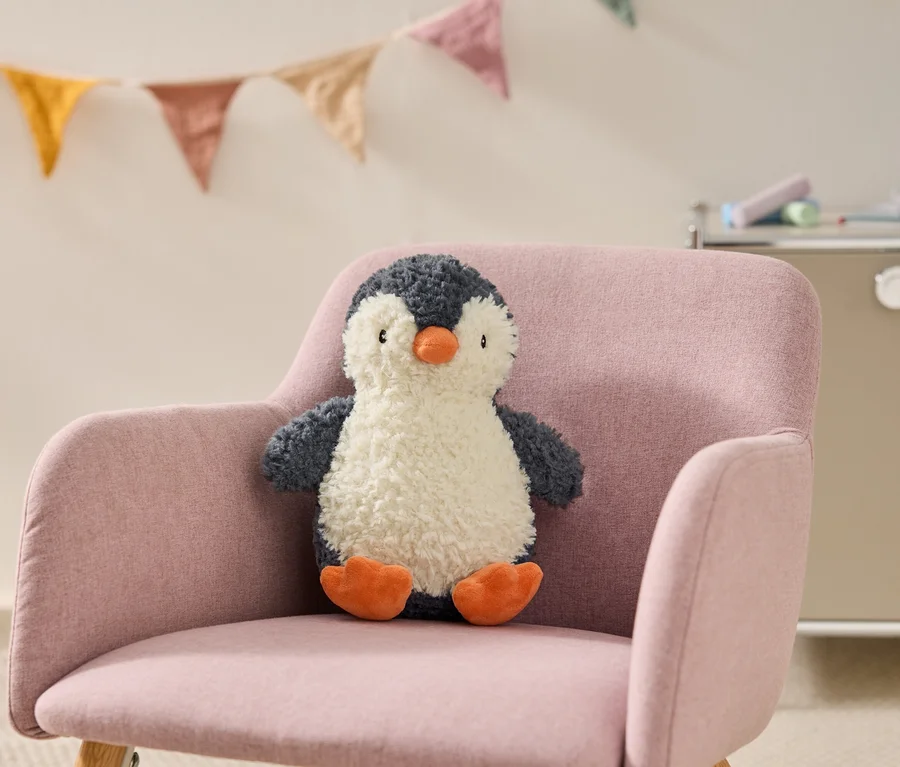 Ein flauschiger dunkelgrau-weißer Pinguin-Plüschtier mit orangefarbenem Schnabel und Füßen sitzt auf einem rosa Sessel. Im Hintergrund hängen bunte Wimpelketten.
