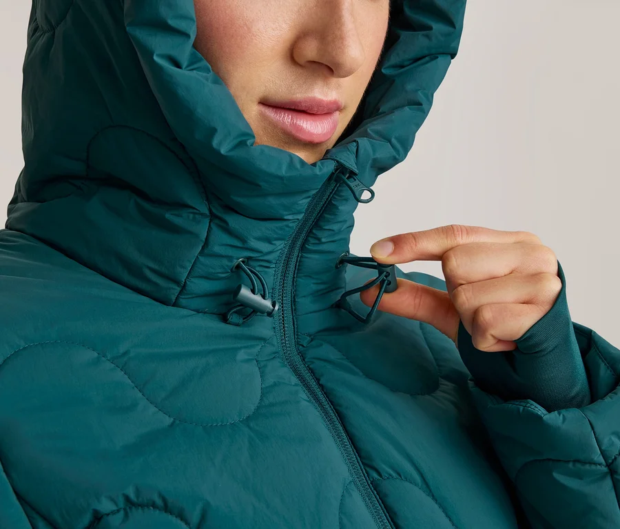 Nahaufnahme einer Frau in einer dunkelgrünen Steppjacke mit Kapuze, die den Kordelzug am Kragen anpasst.
