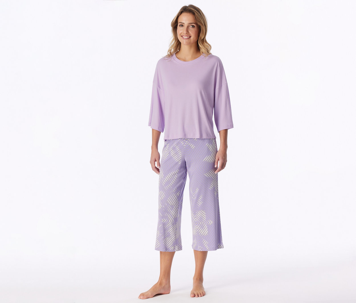 Lächelnde Frau steht barfuß und trägt ein hellviolettes T-Shirt mit Dreiviertelärmeln und passende Caprihosen mit weißem geometrischem Muster.