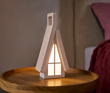 Eine hausförmige Lampe mit hellen Holzseiten und leuchtenden weißen Fenstern steht auf einem runden Holztisch. Im Hintergrund ist ein Bett mit einem rosa Kissen und einer roten Decke zu sehen.