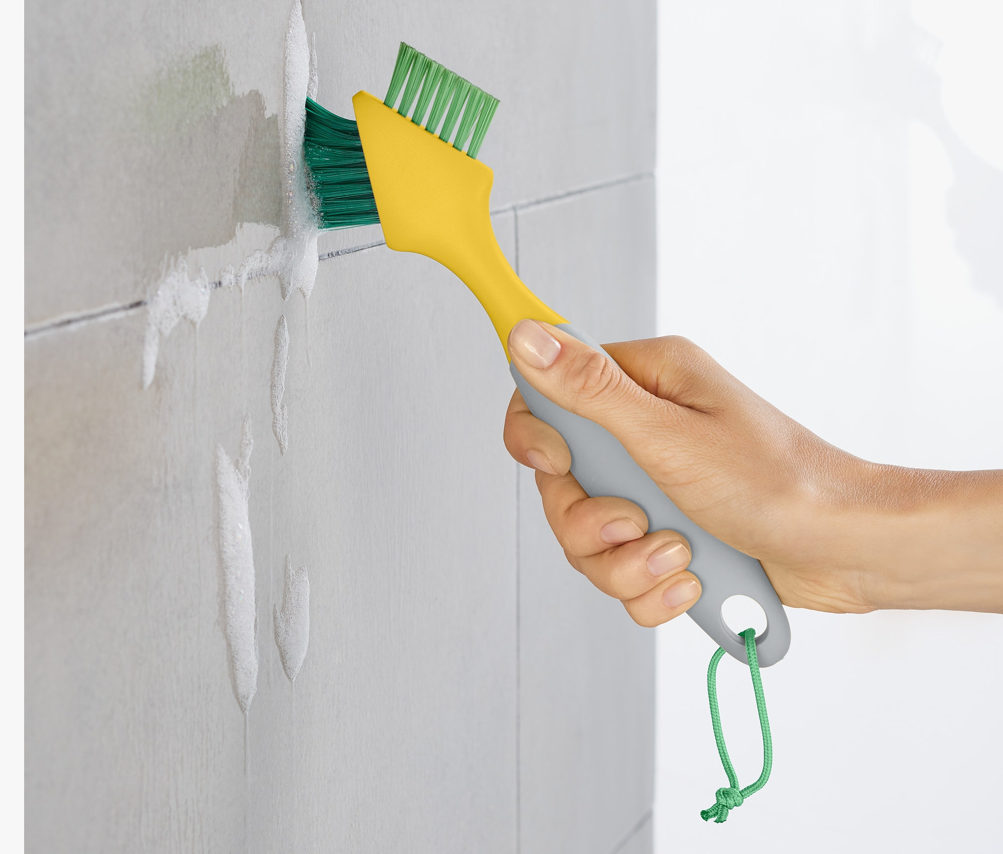 Eine Hand reinigt die Fugen zwischen grauen Fliesen mit einer Fugenbürste.