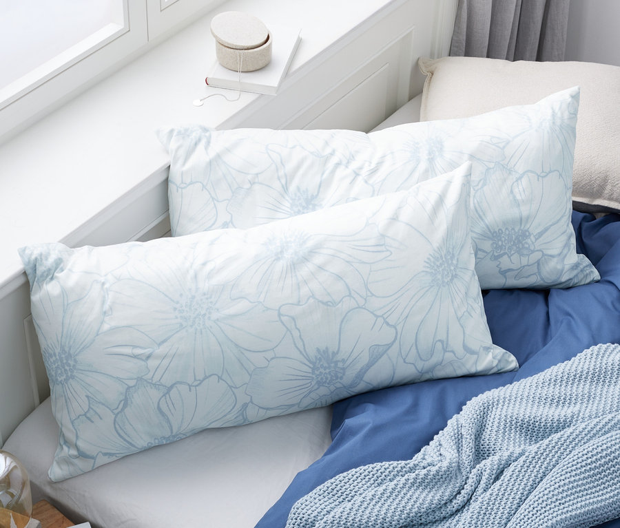 Zwei blau-weiße Renforcé-Kopfkissenbezüge mit floralem Alloverprint liegen auf einem Bett mit blauer Tagesdecke und hellblauer Strickdecke.