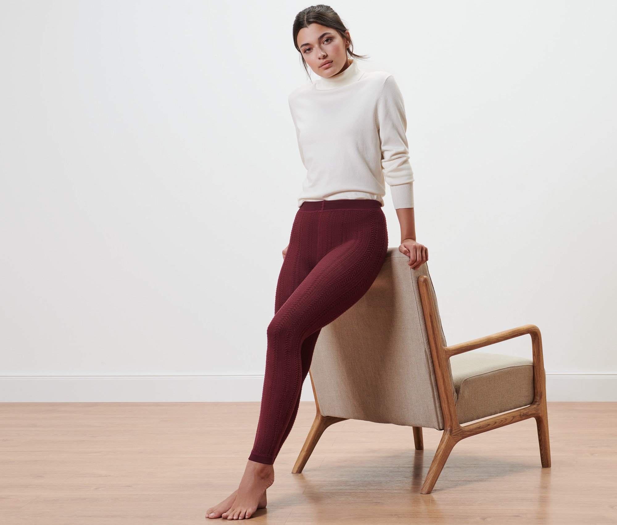 Frau lehnt an Stuhl, trägt Thermoleggings mit Zopfmuster und weißen Rollkragenpullover.