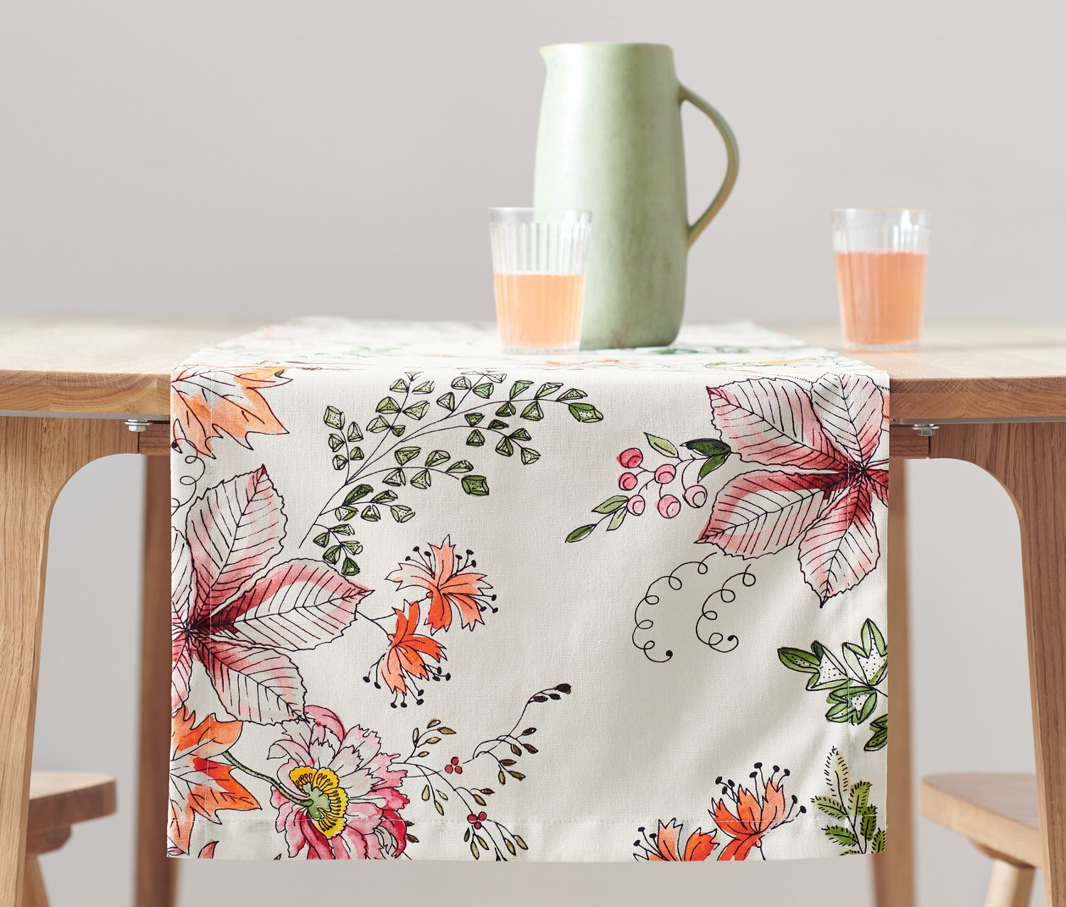 Auf einem Holztisch liegt ein Tischläufer mit Blumenmuster. Auf dem Tisch stehen ein Krug und zwei Gläser mit orangefarbener Flüssigkeit.