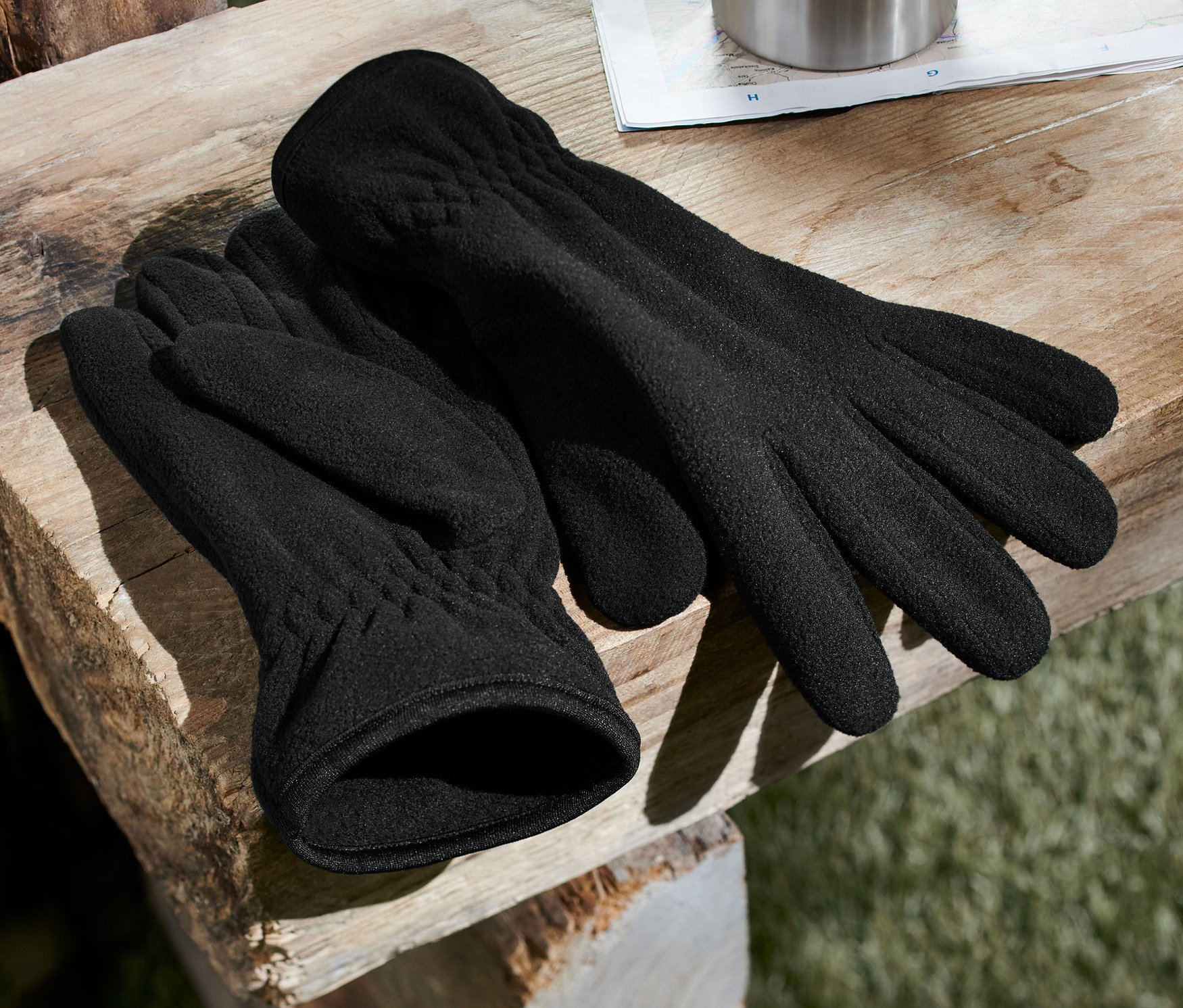 Schwarze Fleece-Handschuhe liegen auf einer Holzbank neben einem Rucksack und einer Thermoskanne.