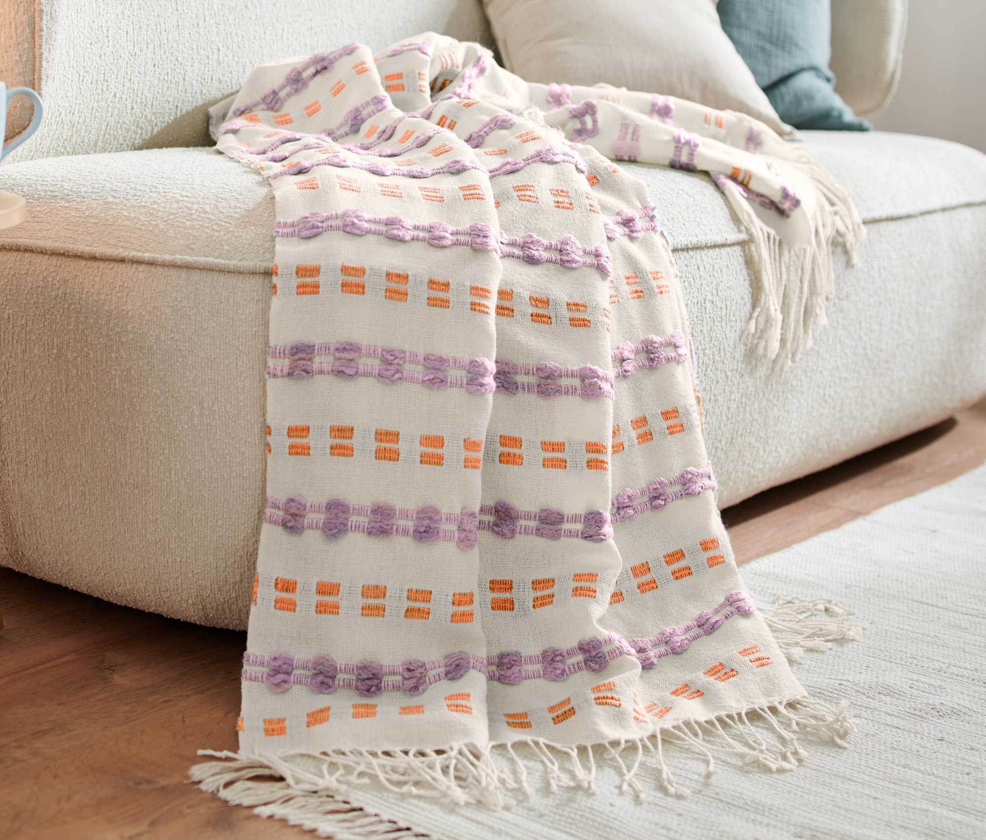 Auf einem beigen Sofa liegt eine Wohndecke mit lila- und orangefarbenen Details.