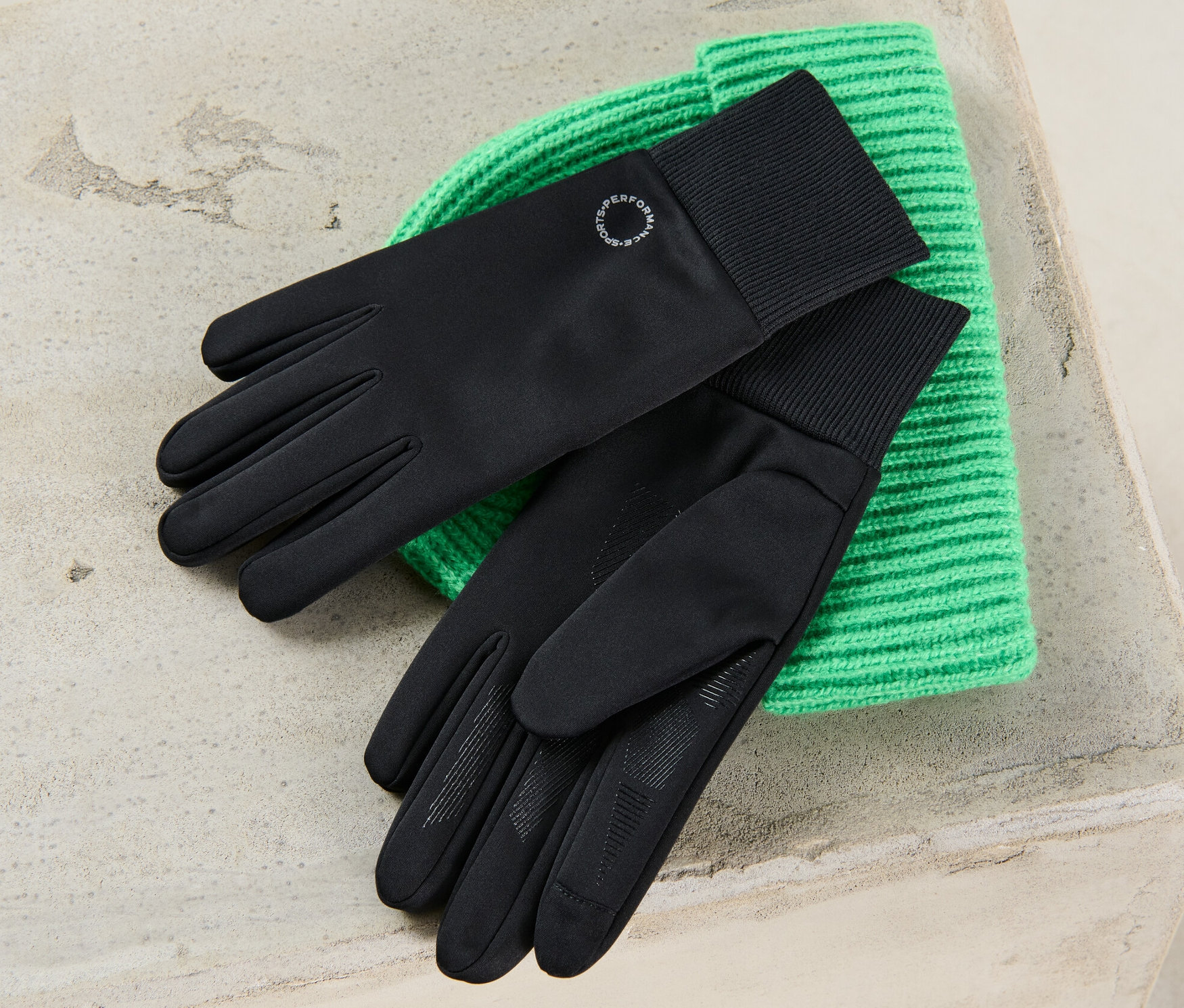 Schwarze Windprotection-Handschuhe liegen auf einer grünen Strickmütze.