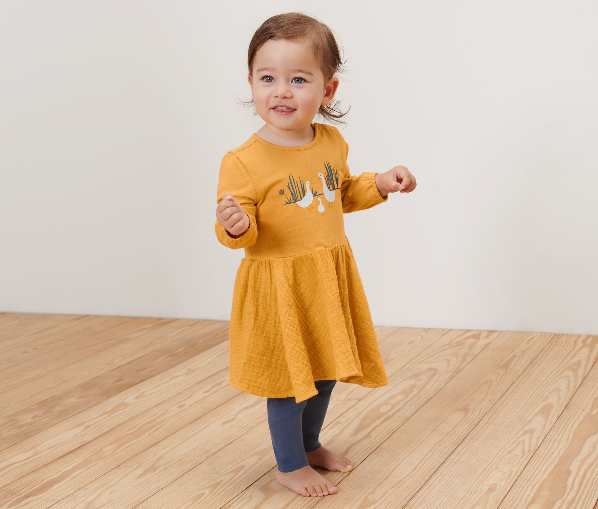 Ein lächelndes kleines Mädchen steht auf einem Holzboden und trägt ein gelbes Baby-Webkleid.