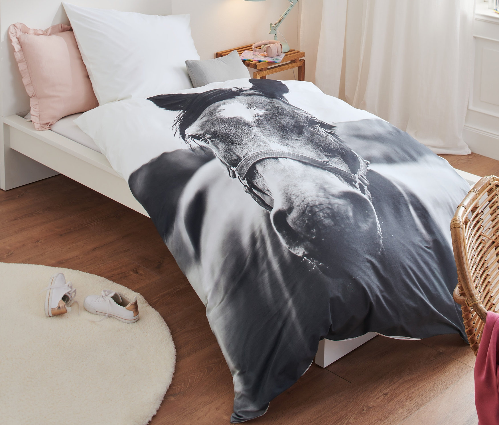 Ein Bett mit Perkal-Pferde-Bettwäsche, Normalgröße. Auf dem Boden liegt ein runder Teppich und neben dem Bett steht ein Korbsessel.