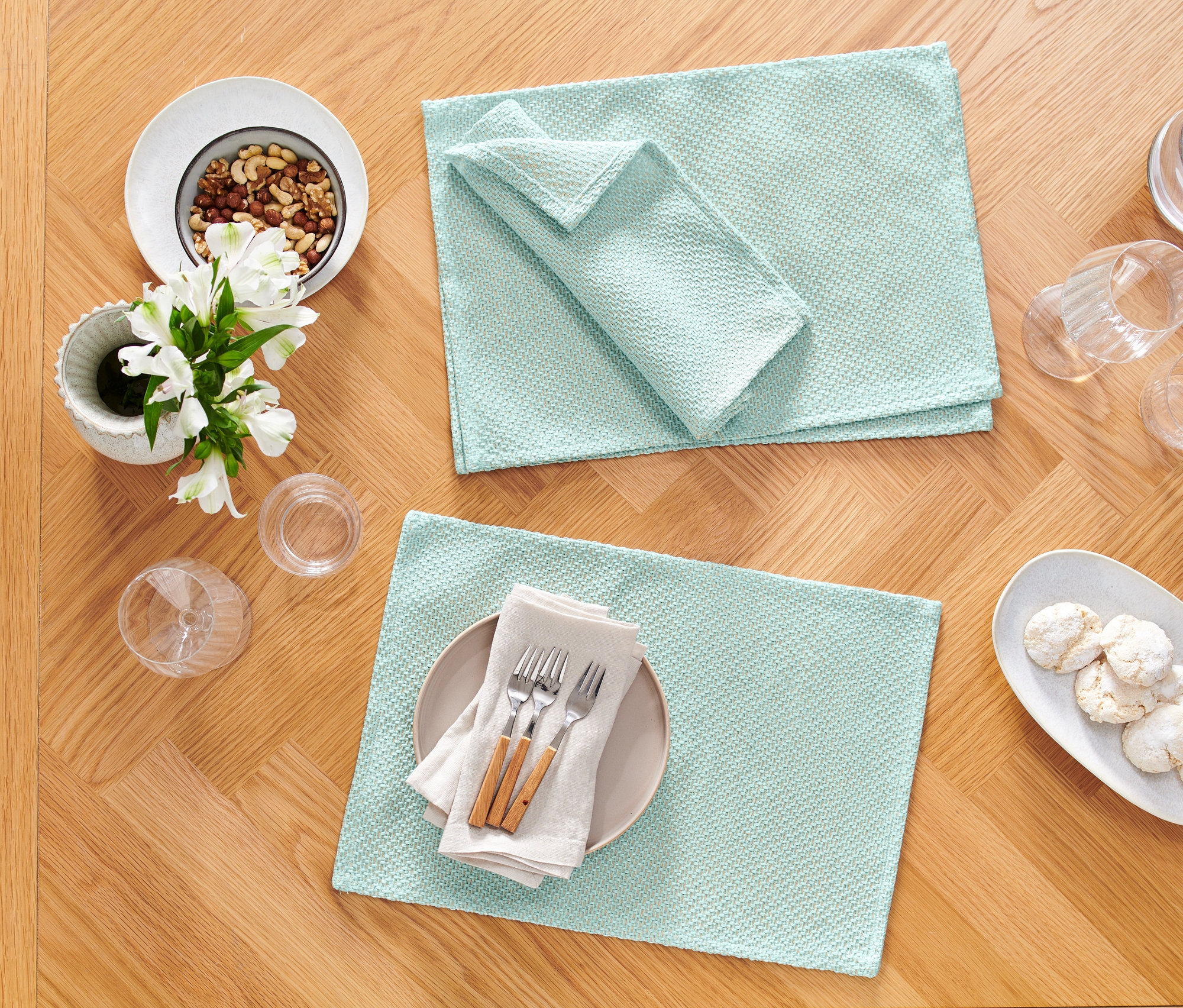 Zwei hellblaue Platzsets und eine passende Serviette liegen auf einem Holztisch neben einem Teller mit Nüssen, Blumen und Gläsern.