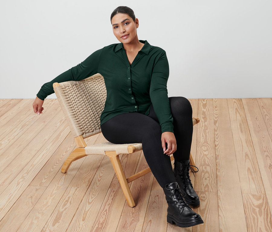 Eine Frau sitzt auf einem Korbstuhl und trägt ein dunkelgrünes Blusenshirt mit Knopfleiste, Thermo-Leggings mit Prägung und Leder-Boots.