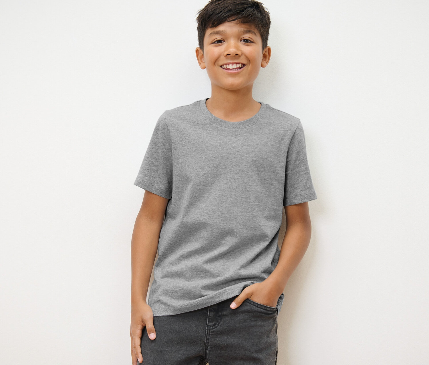 Ein Junge steht und trägt ein graues T-Shirt und Jeans. Er trägt 3 Kinder-T-Shirts in Schwarz, Grau und Weiß.