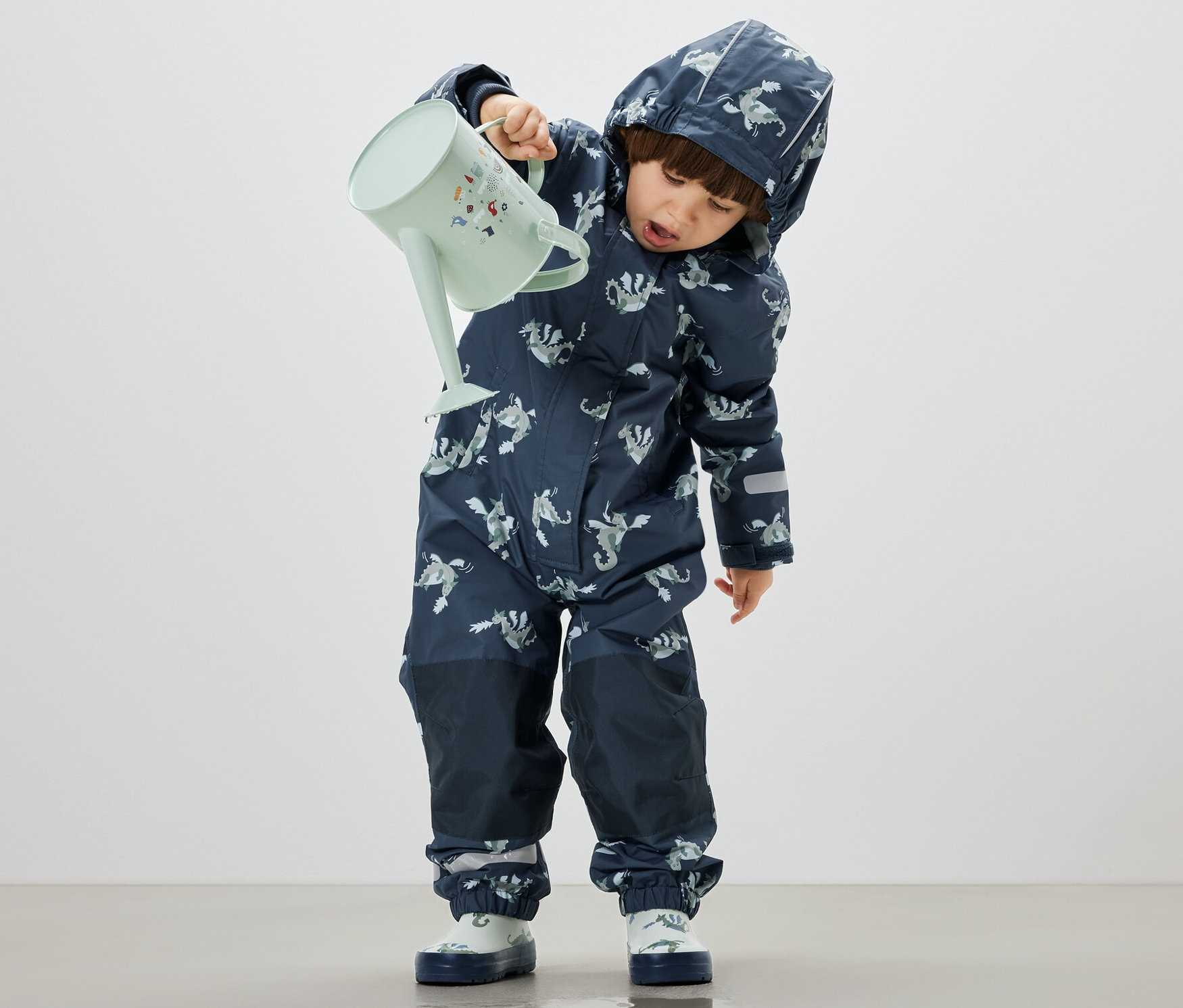 Junge gießt Wasser aus einer pastellgrünen Gießkanne auf den Boden, trägt dabei einen Kinder-Regen-Einteiler, Ritter.