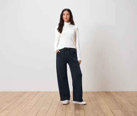 Frau steht mit Händen in den Taschen einer dunkelblauen, weiten Hose, einem weißen Rollkragenpullover und weißen Turnschuhen.