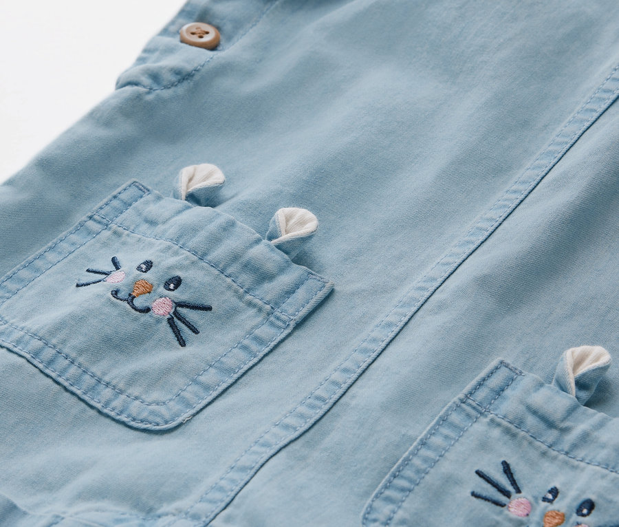 Detail einer Kinder Chambray-Latzhose mit Hasenstickerei auf der Tasche.