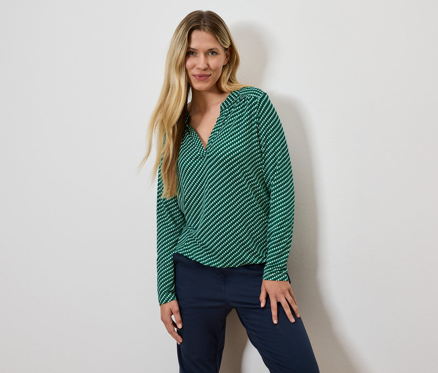 Frau mit blonden Haaren posiert in einer grün gestreiften Bluse und dunkelblauen Hosen.