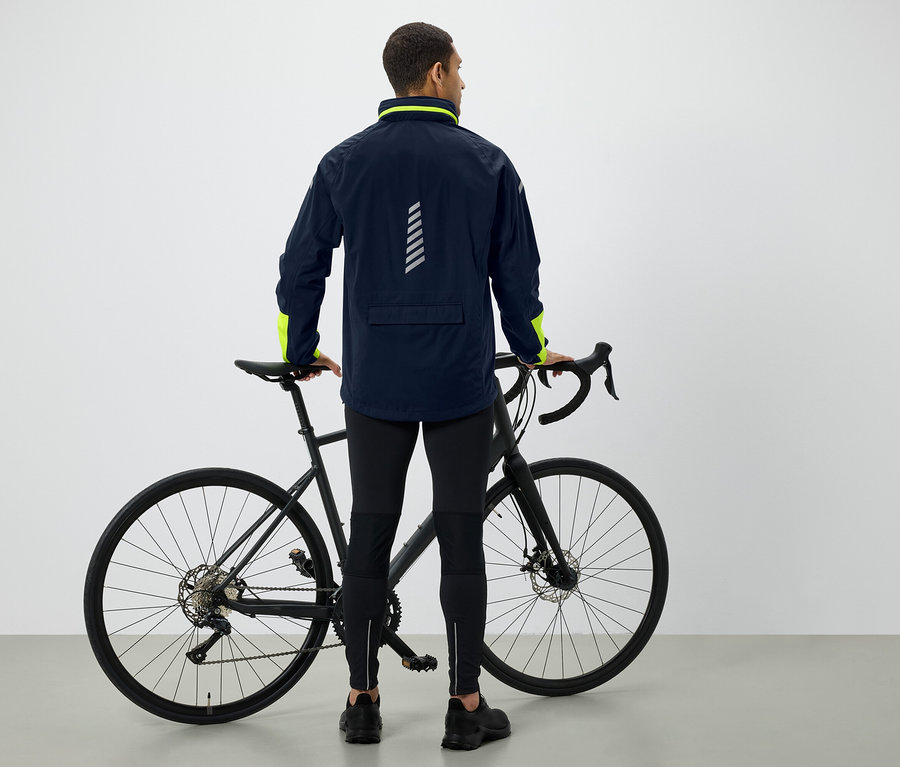 Ein Mann steht mit dem Rücken zur Kamera und hält ein schwarzes Fahrrad. Er trägt eine dunkelblaue Jacke mit neongrünen Details und eine schwarze Hose.